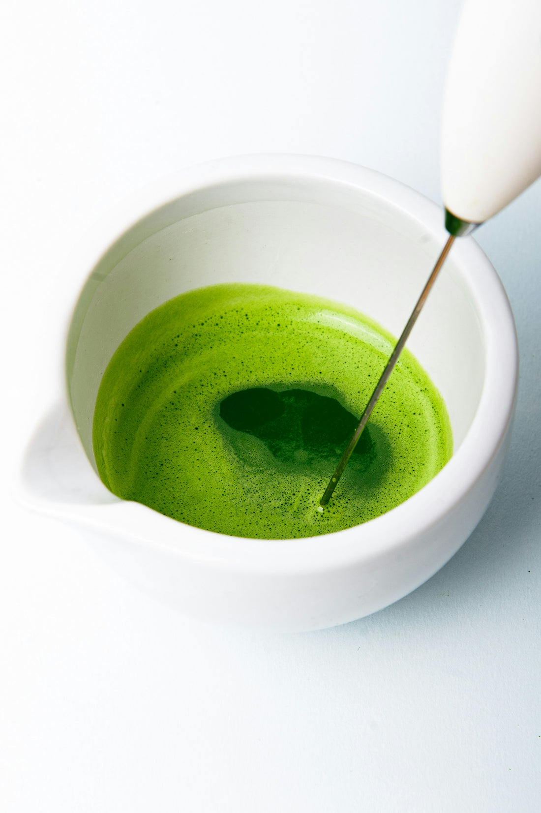 A white bowl filled with dark green matcha being whisked with a bamboo whisk, set against a clean white background.