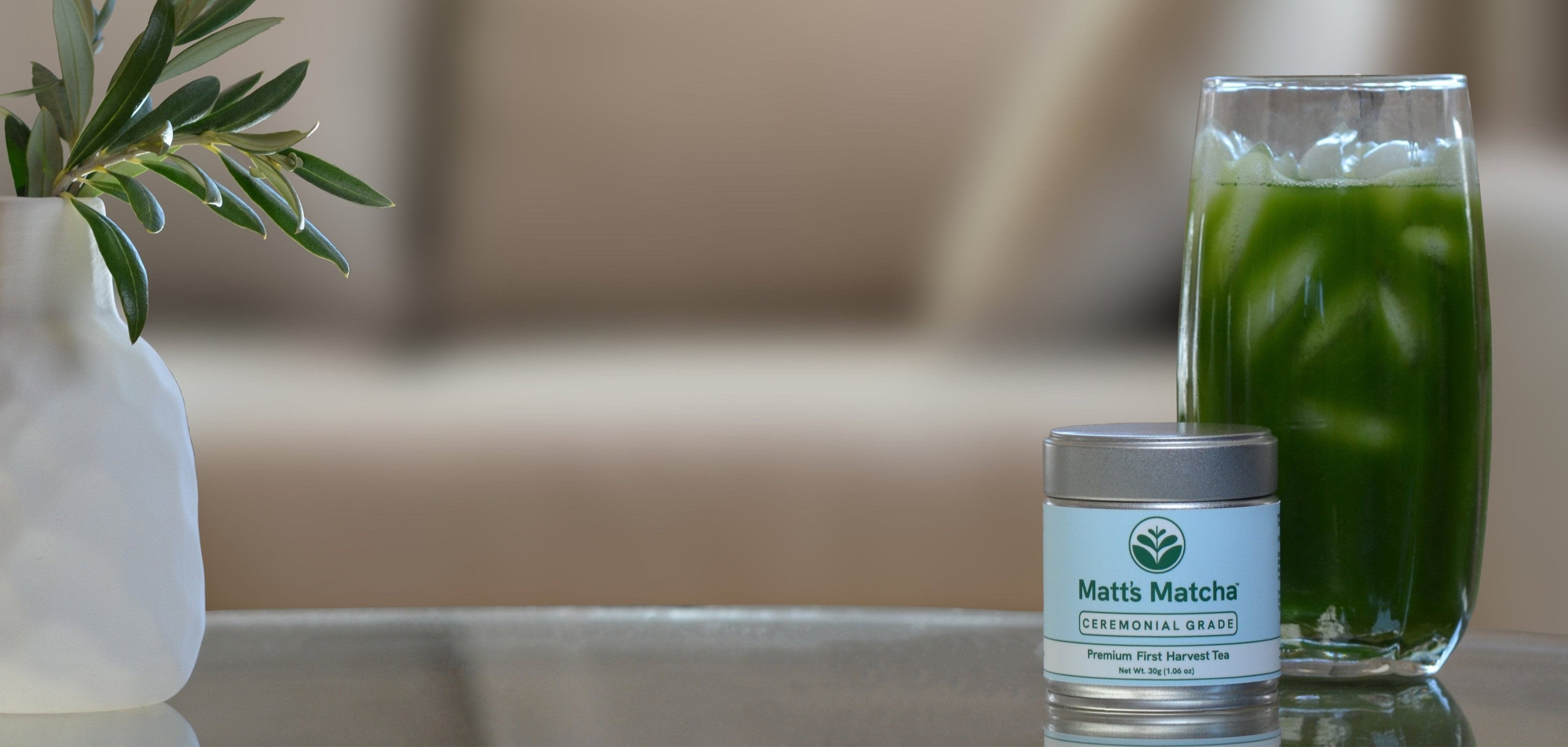 A plant in a white vase sits next to a metal tin of matcha with a blue label and green 'Matt's Matcha' lettering. Behind the tin is a glass of green iced matcha tea. The items are arranged on a table, with a couch visible in the background.