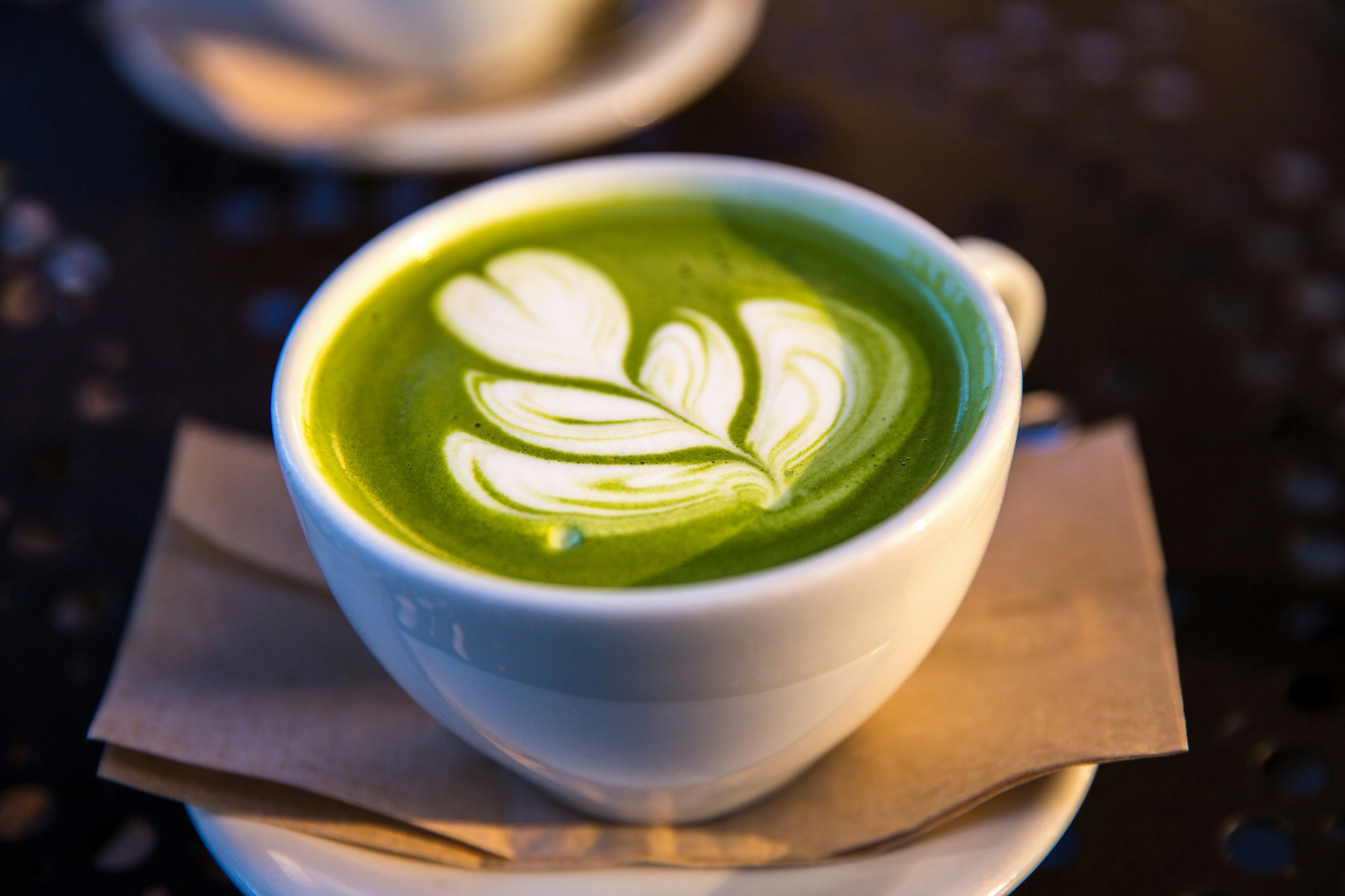 A hot green matcha latte with intricate latte art in a white mug set against a cafe table background.